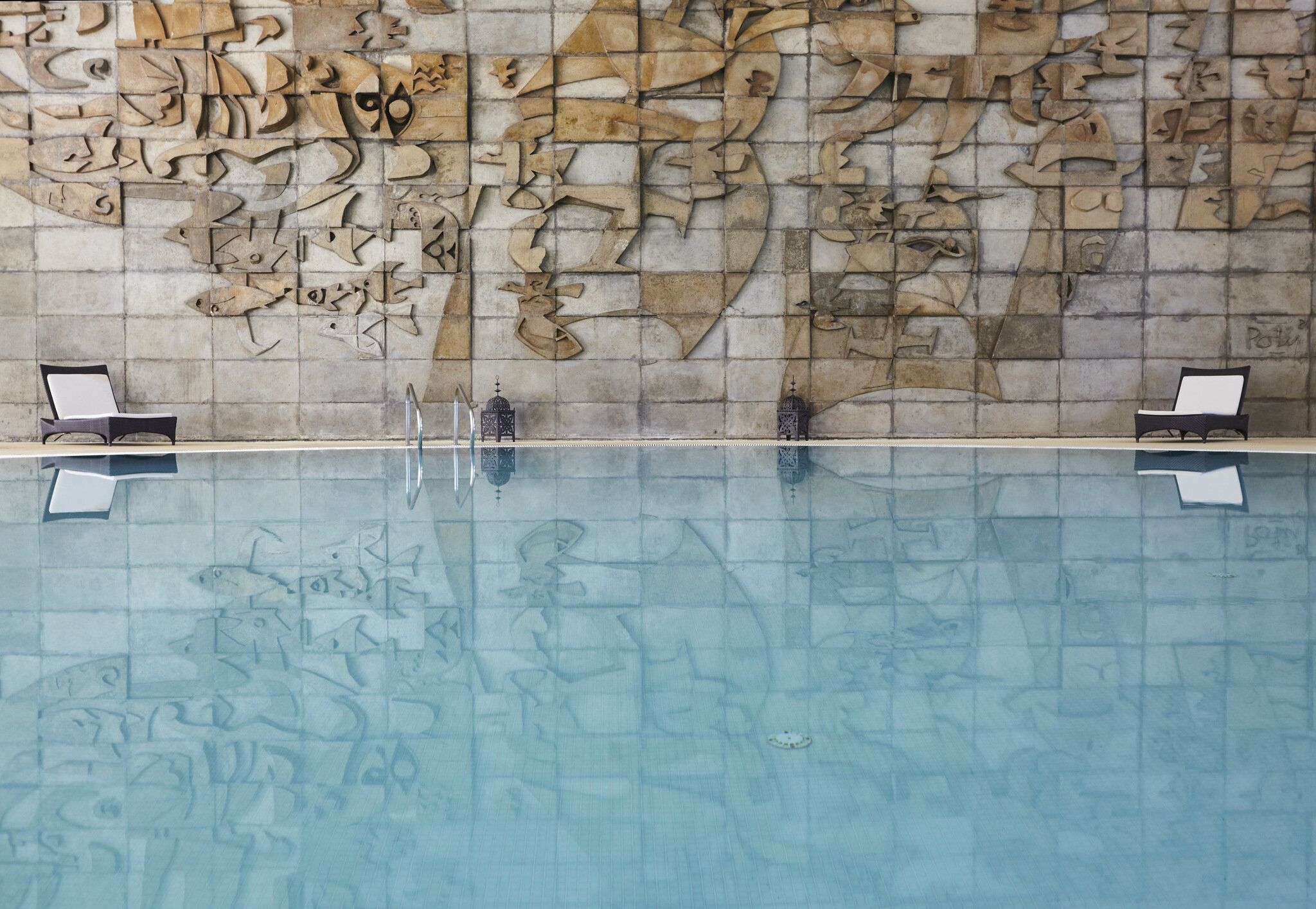 Una piscina con un muro de azulejos con diseños abstractos. Hay dos tumbonas a cada lado de la piscina.