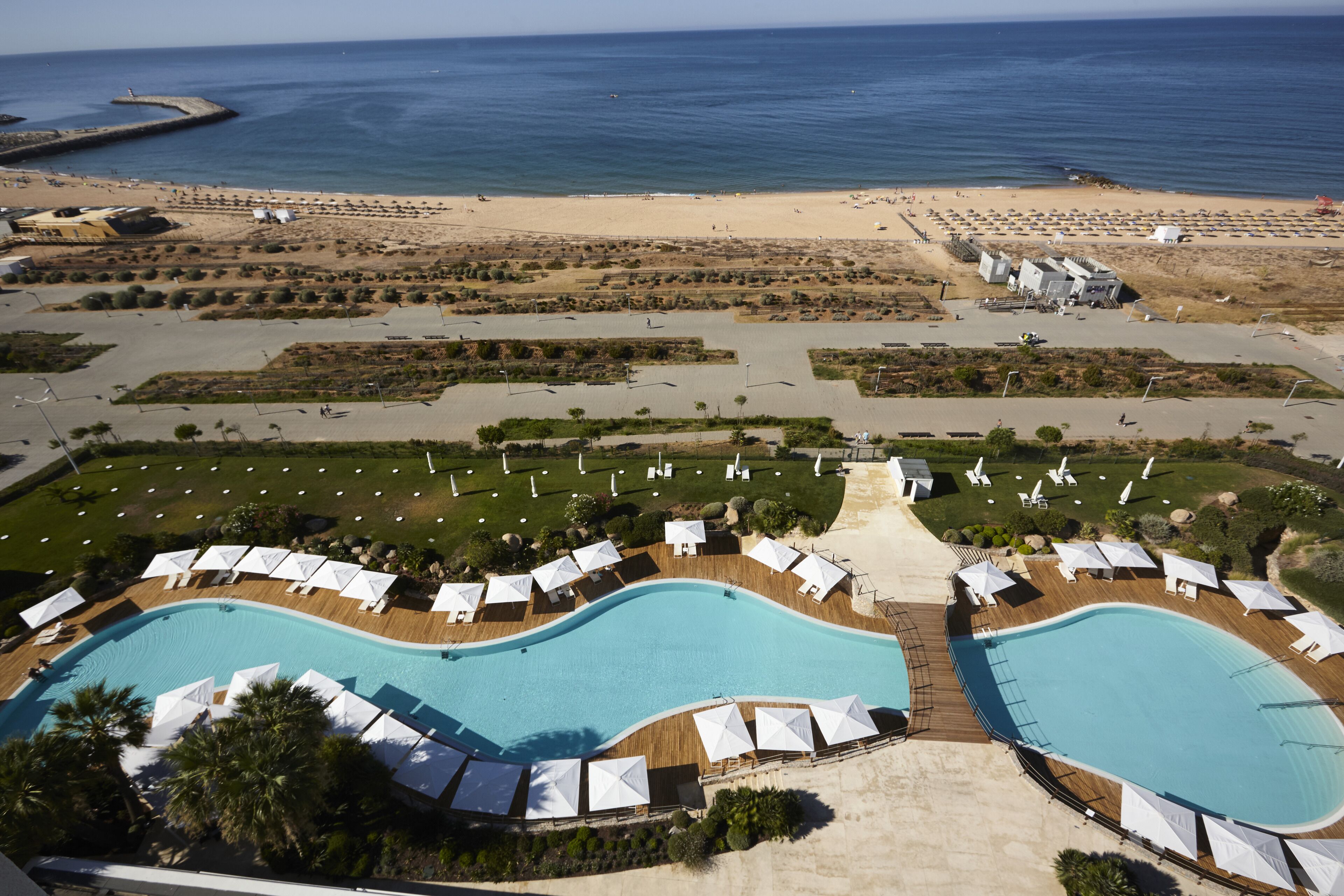Vista aérea de un resort con dos piscinas grandes, tumbonas y una zona de playa.
