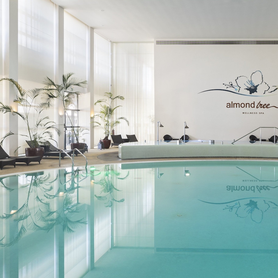 A modern indoor pool area with a logo for 'almond tree wellness spa' on the wall, surrounded by lounge chairs and potted plants.