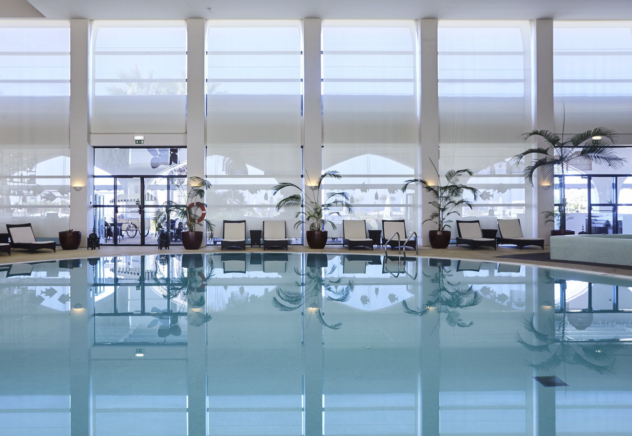 A large indoor swimming pool with clear glass walls, multiple lounge chairs, and potted plants.