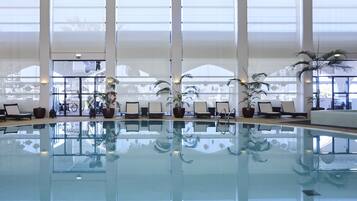 A large indoor swimming pool with clear glass walls, multiple lounge chairs, and potted plants.