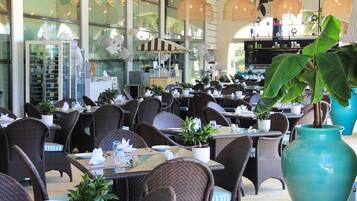A restaurant with wicker furniture, tables set with white napkins, and large potted plants.