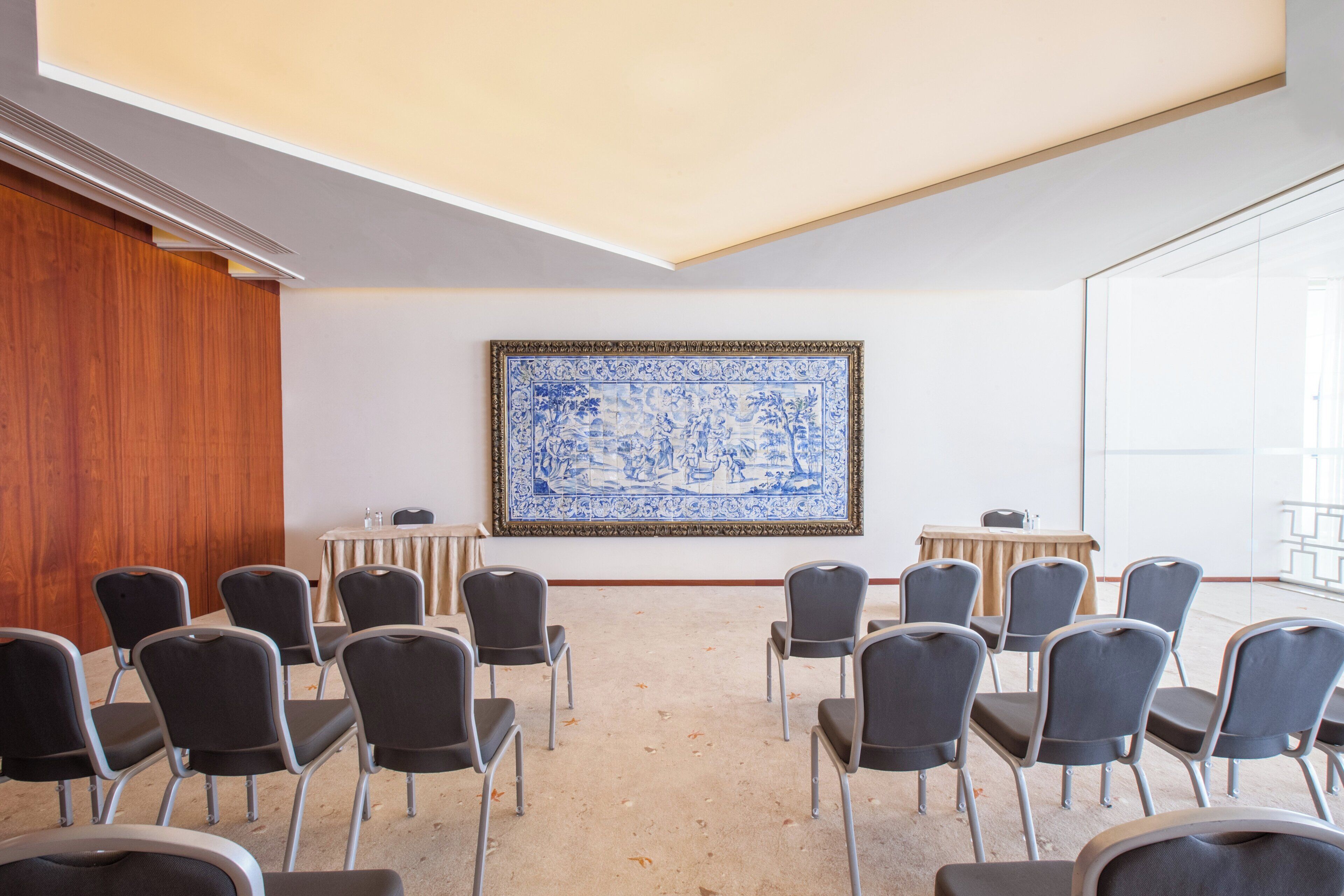 Un salón de conferencias con techo alto, una gran obra de arte de azulejos azules y blancos en la pared, y filas de sillas frente a un atril.