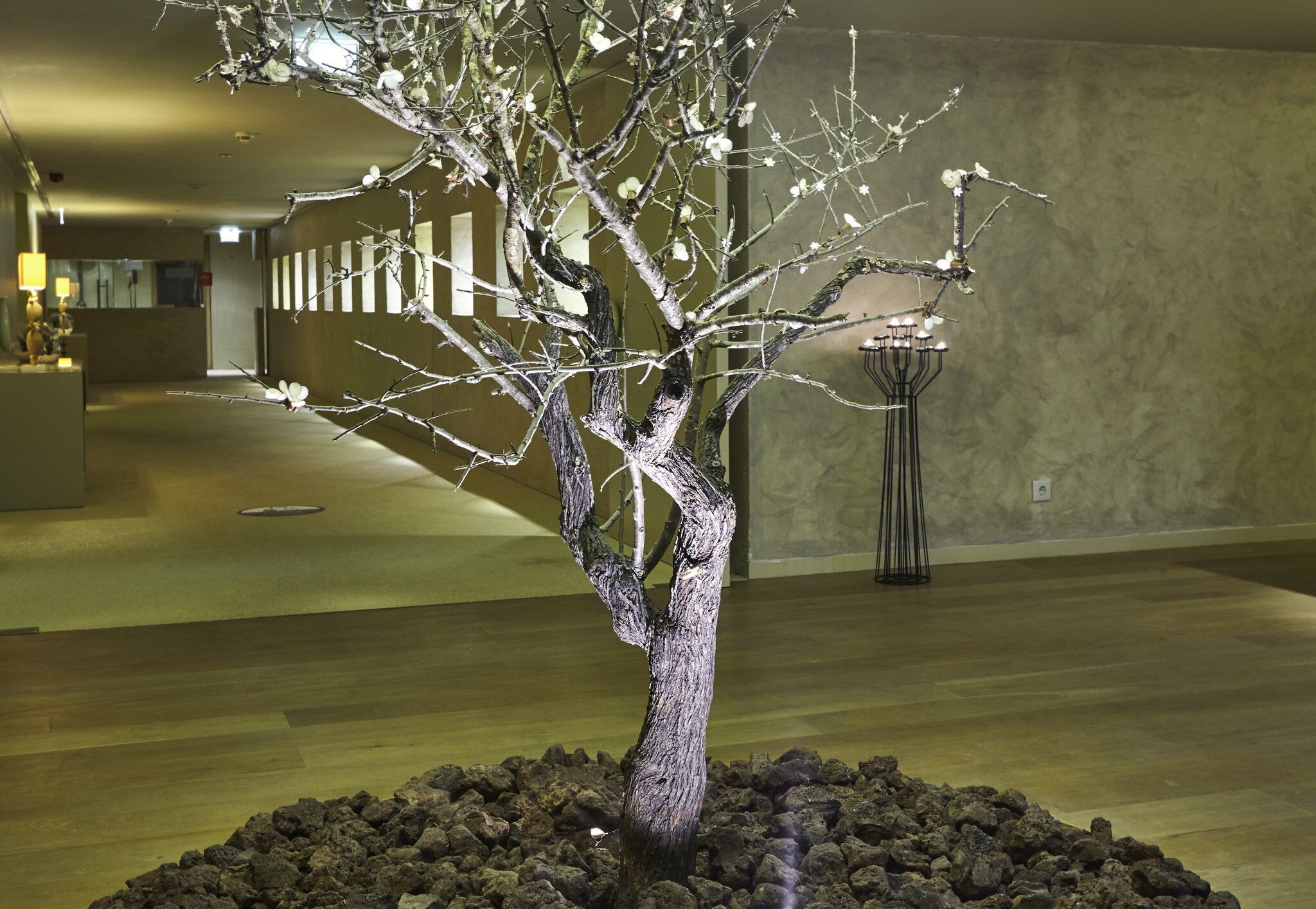 Une sculpture d’arbre, avec ses branches et ses feuilles, est placée sur un lit de pierres dans une pièce dont le sol est en bois et dont un mur est orné de lignes verticales.