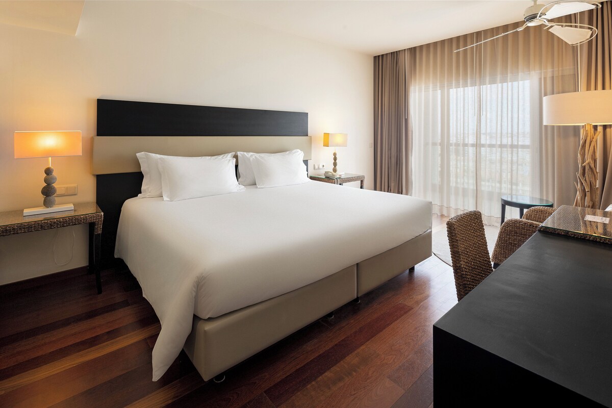 A hotel room with a large bed, two bedside tables with lamps, a desk with a chair, and a large window with curtains.