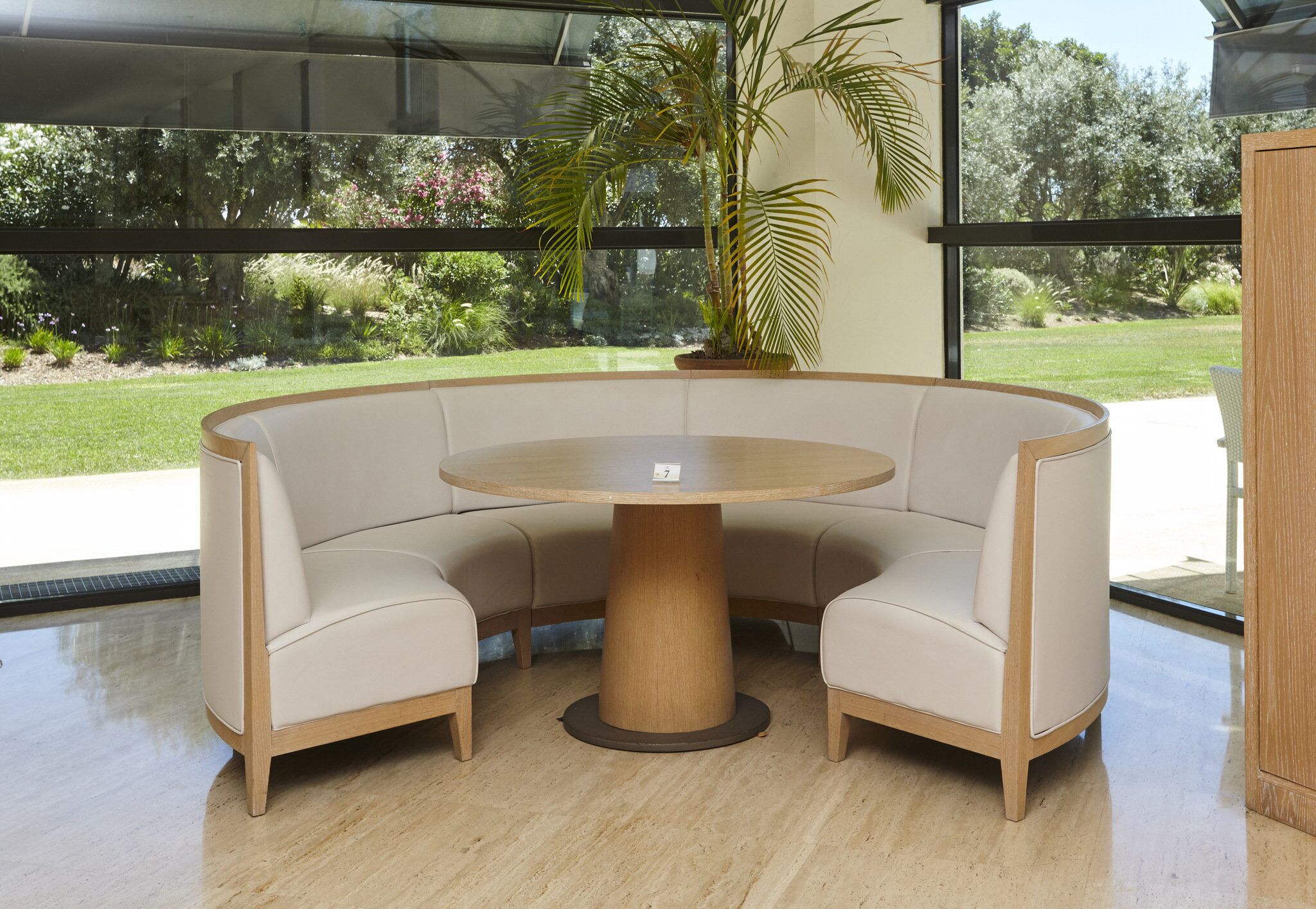 A round table with a wooden base, surrounded by beige upholstered chairs, in a room with large windows and a view of greenery outside.