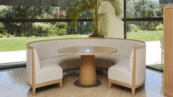 A round table with a wooden base, surrounded by beige upholstered chairs, in a room with large windows and a view of greenery outside.