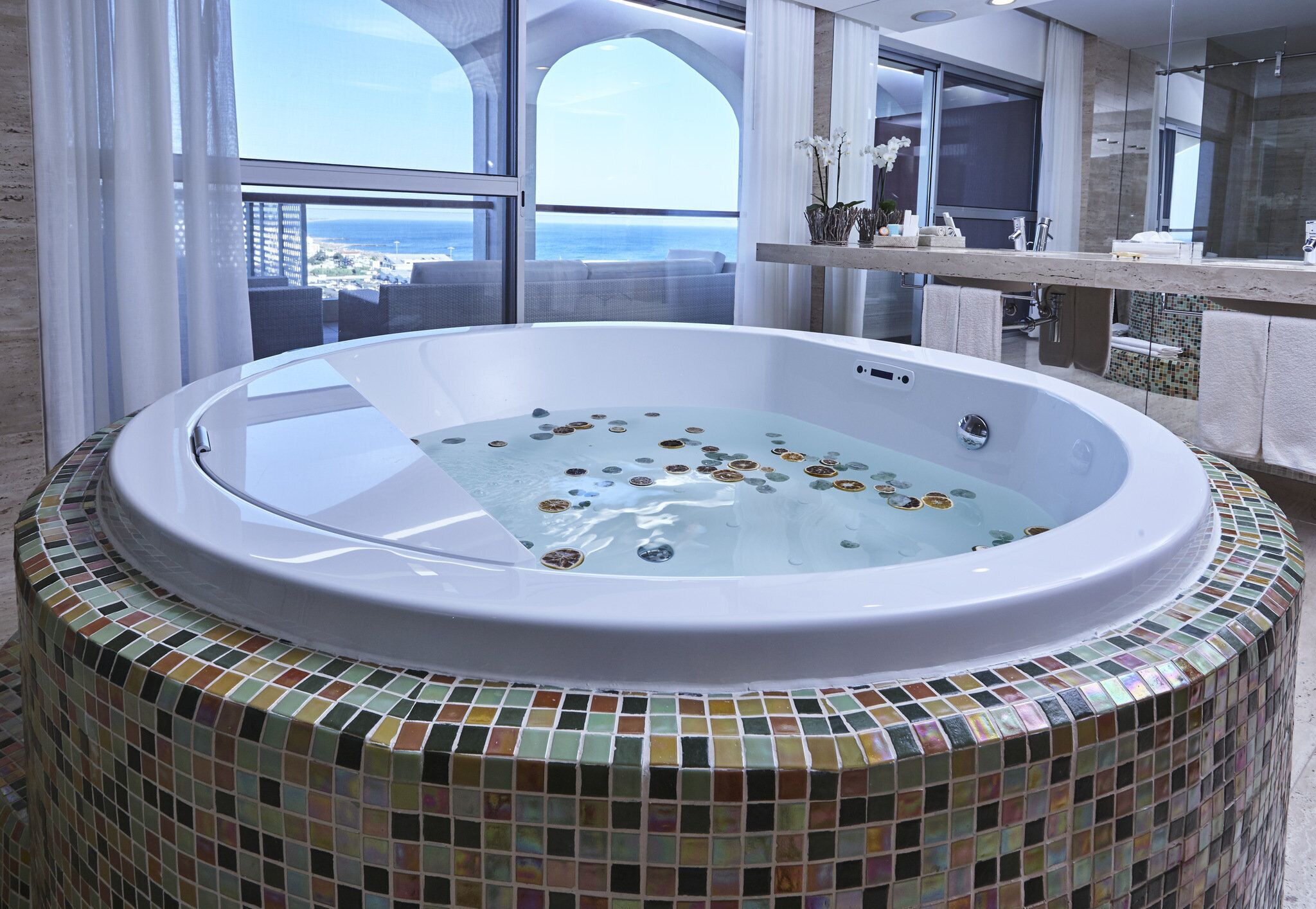 A hot tub with a mosaic tile surround, a large window with a view of the sea, and a modern bathroom interior.