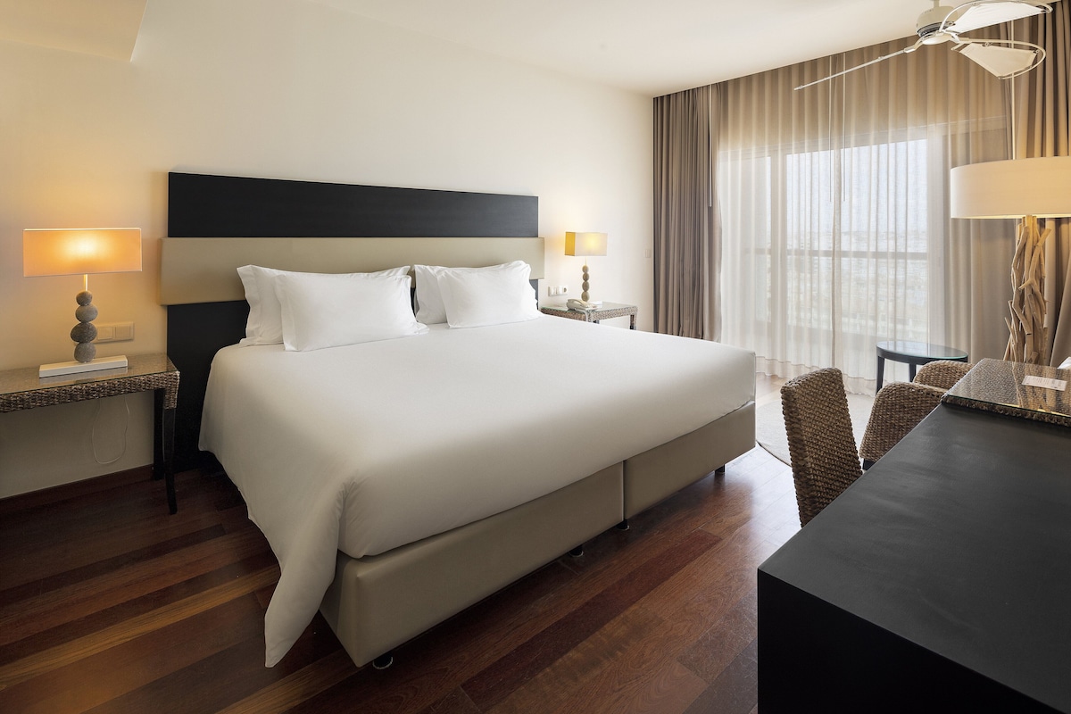 Una habitación de hotel con una cama grande, dos mesitas de noche con lámparas, un escritorio con silla y un ventanal con cortinas.