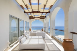 Un balcone baciato dal sole con sedute imbottite bianche, ampie finestre e vista sull'oceano.