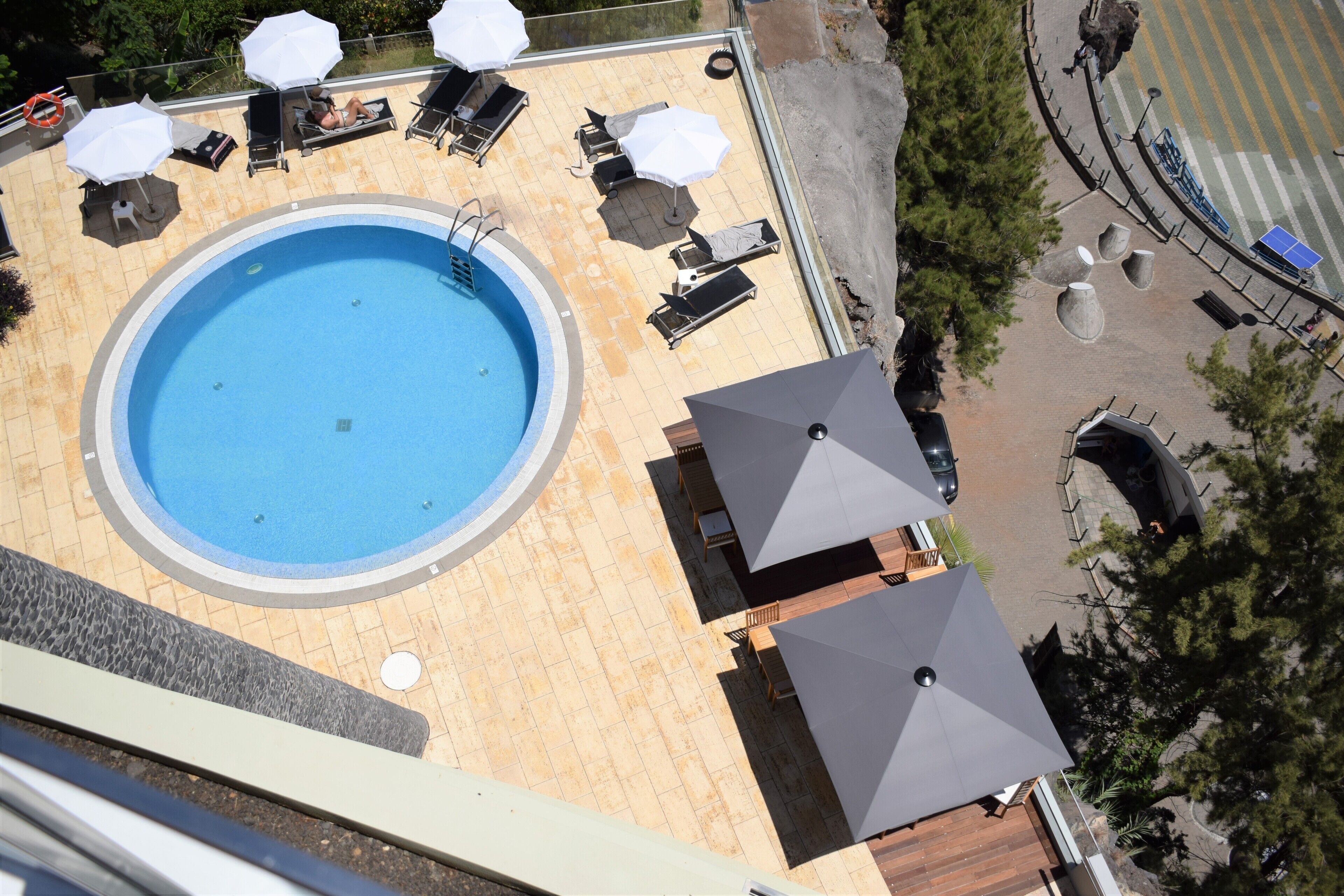 En pool på taget med liggestole og parasoller, omgivet af en belagt terrasse og træer.