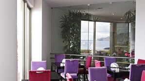 A dining area with purple chairs and a table, large windows with a view of the sea and mountains, and a potted plant. - Madeira Regency Cliff (Funchal)