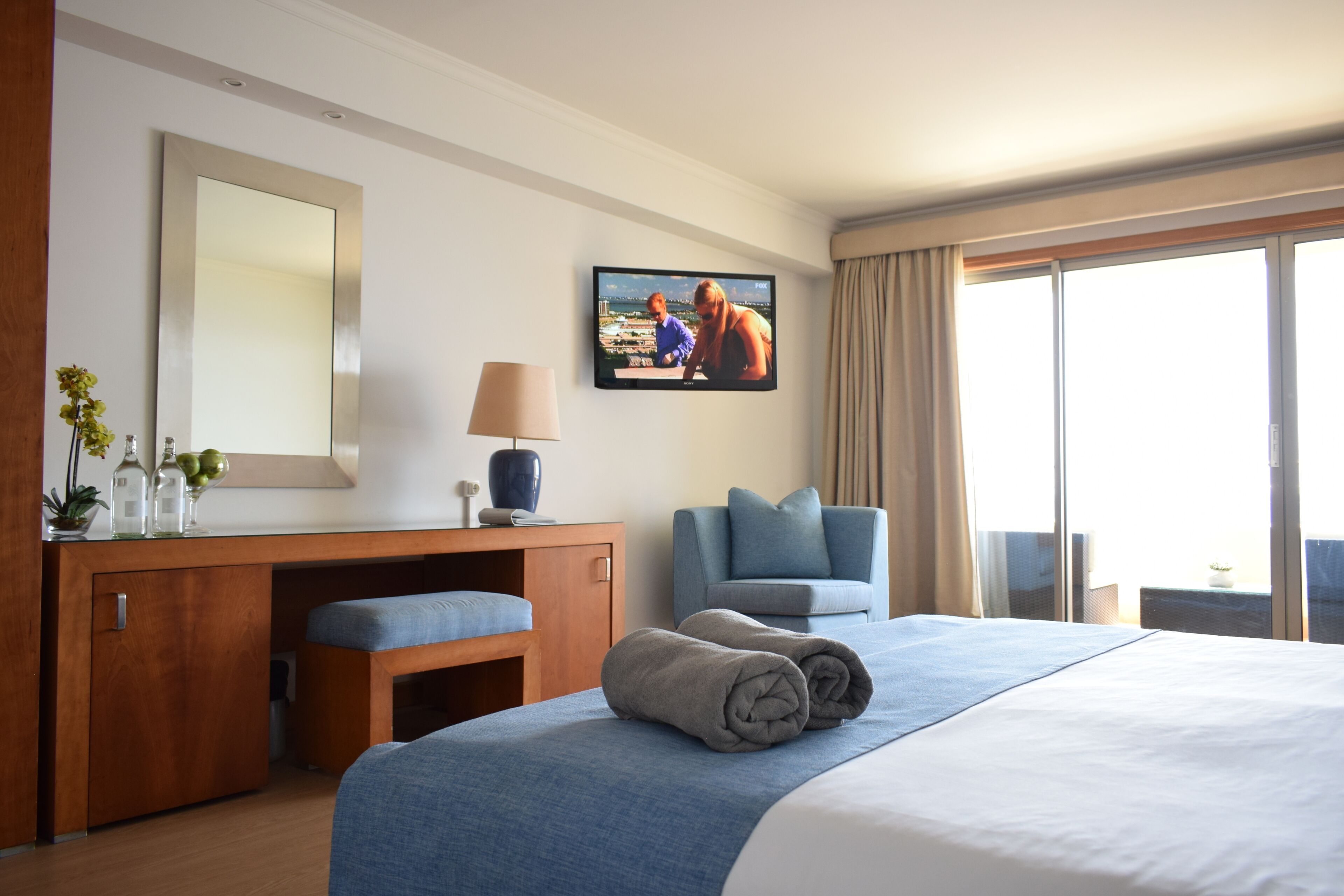 Ein Hotelzimmer mit einem großen Bett, einem Schreibtisch mit Lampe, einem Sessel, einem an der Wand befestigten Fernseher und einem Fenster mit Vorhängen.