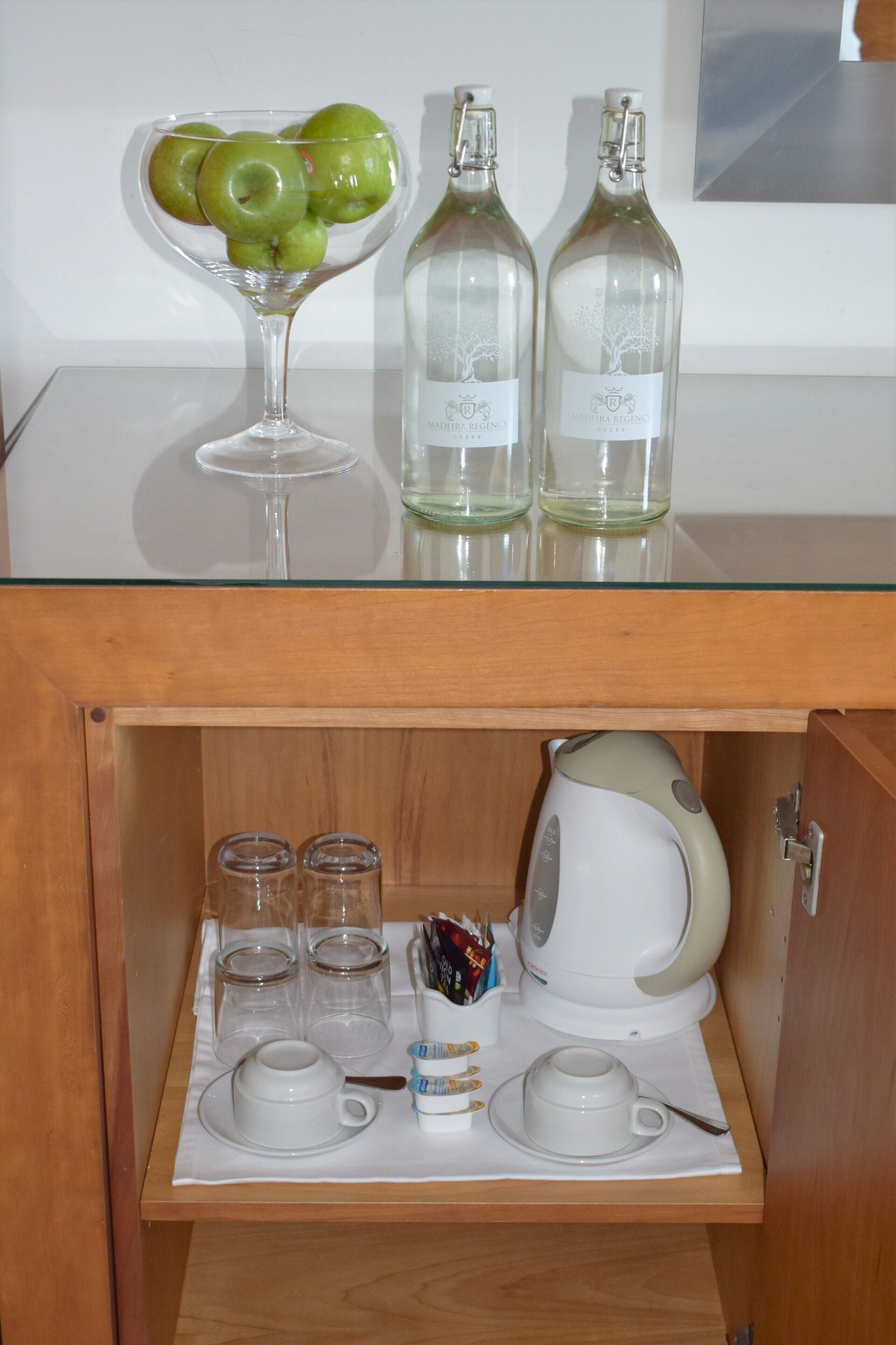 A glass table with a bowl of green apples, two bottles, and a wine glass. Below, a wooden cabinet contains a tea kettle, cups, and sugar packets.