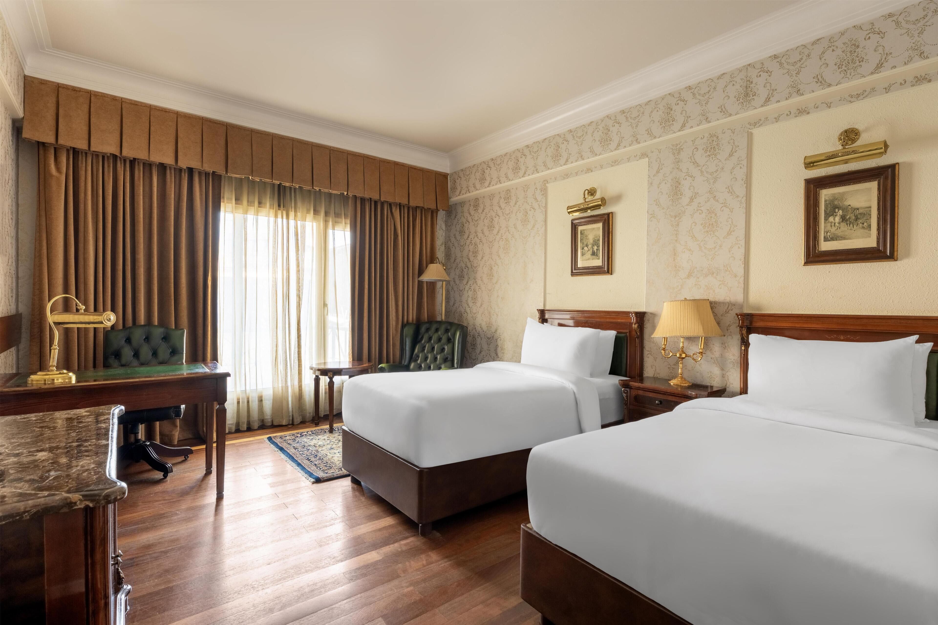 A hotel room with two beds, a desk, a chair, and a large window with curtains.