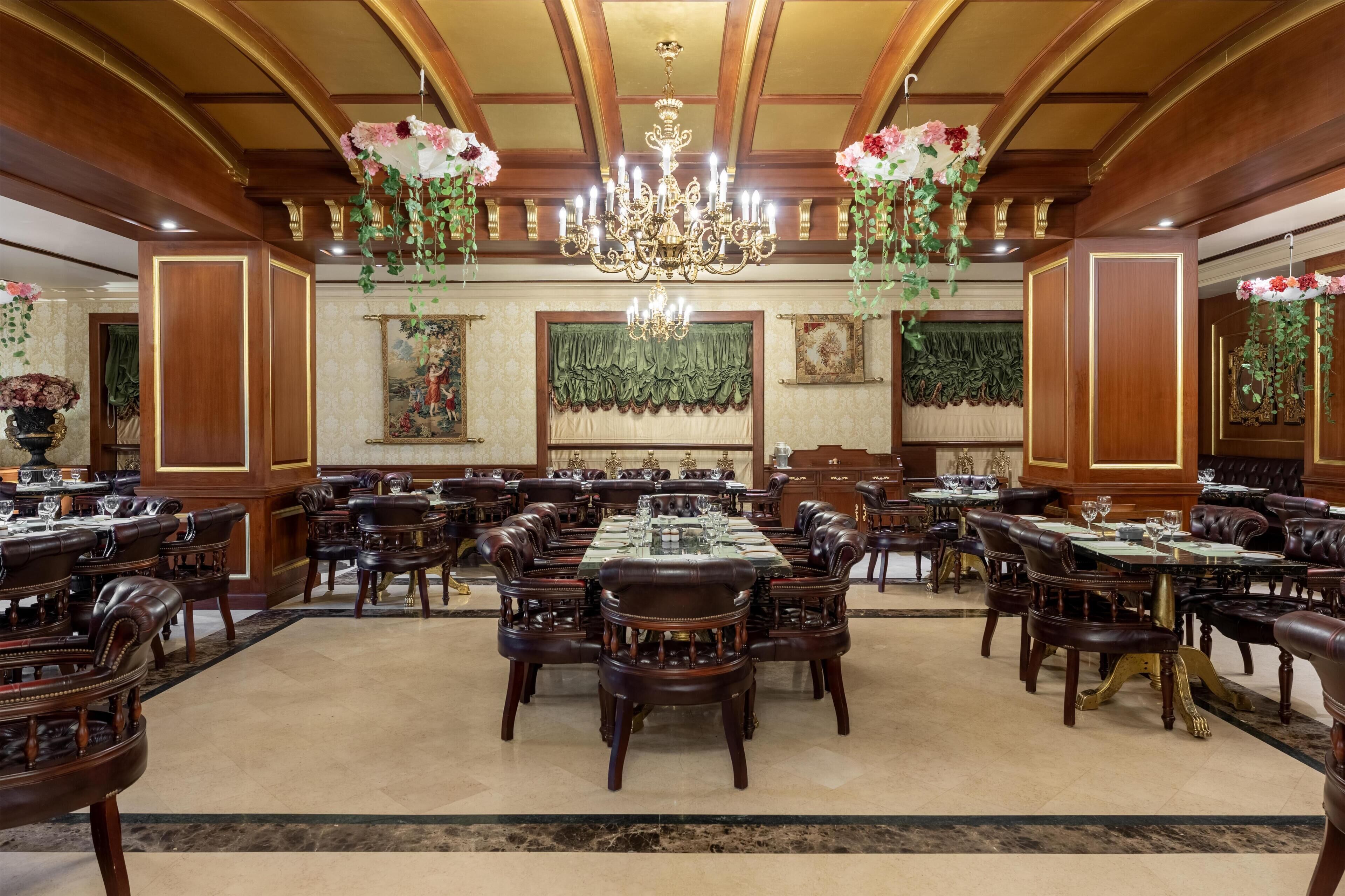 A grand dining hall with multiple round tables and leather-upholstered chairs, ornate chandeliers, and floral decorations.