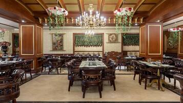 A grand dining hall with multiple round tables and leather-upholstered chairs, ornate chandeliers, and floral decorations.