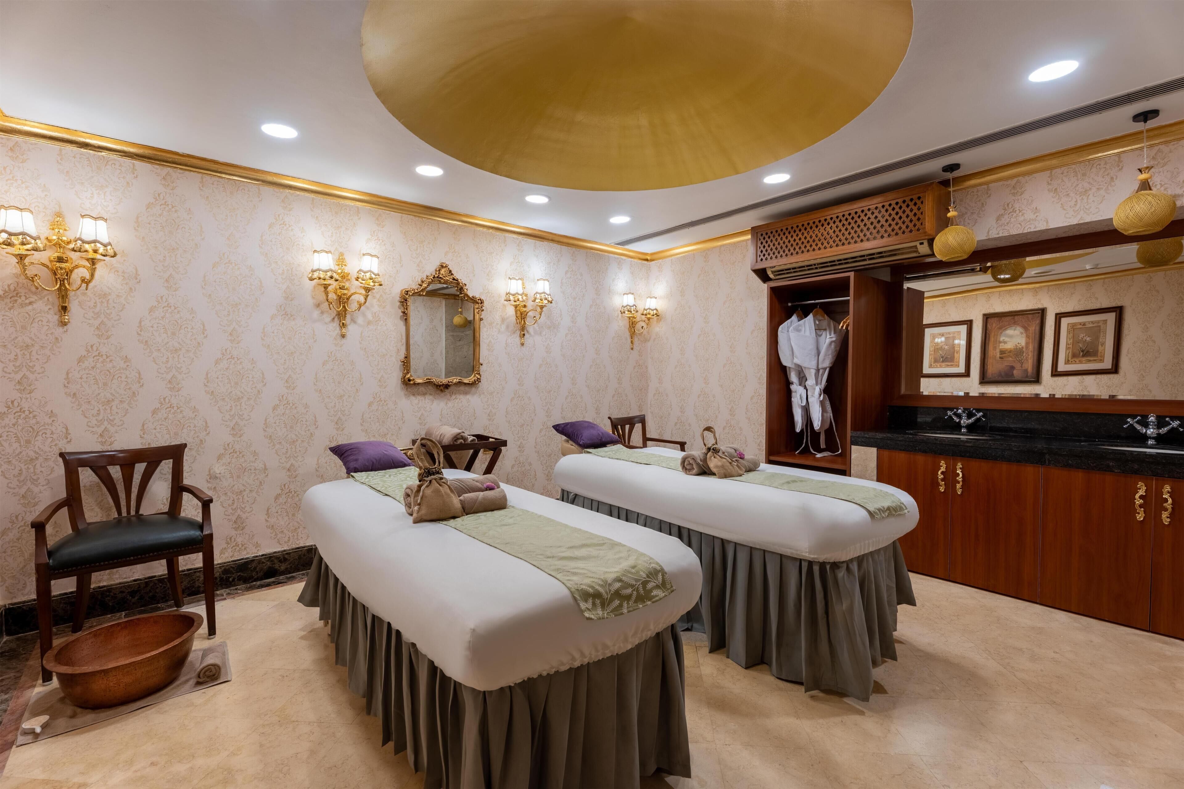 A spa room with two massage tables, a wooden chair, a bathtub, and a vanity with a mirror and lights.