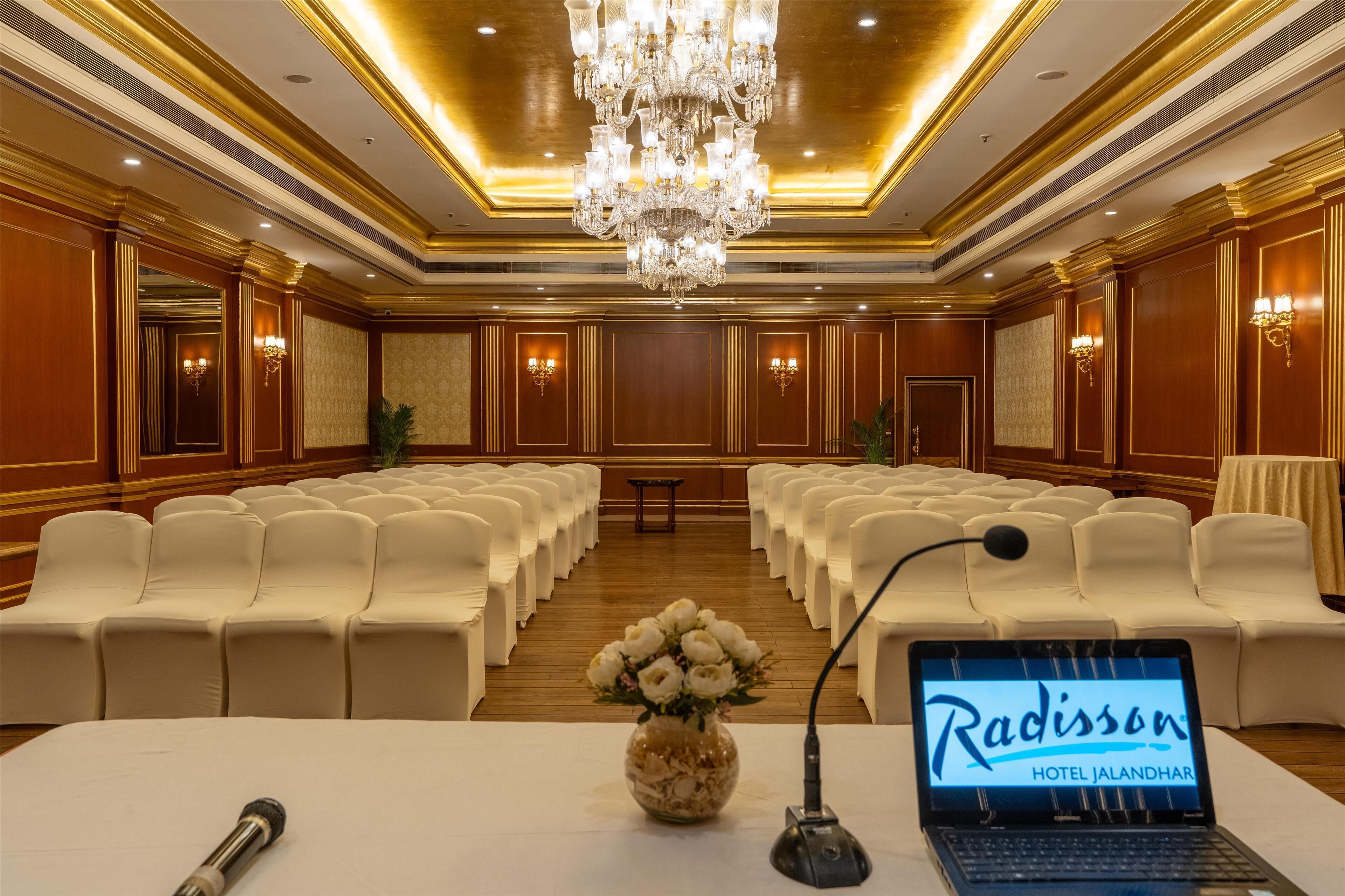 A conference room with rows of white chairs, a chandelier, and a laptop displaying 'Radisson Hotel Jalandhar'.
