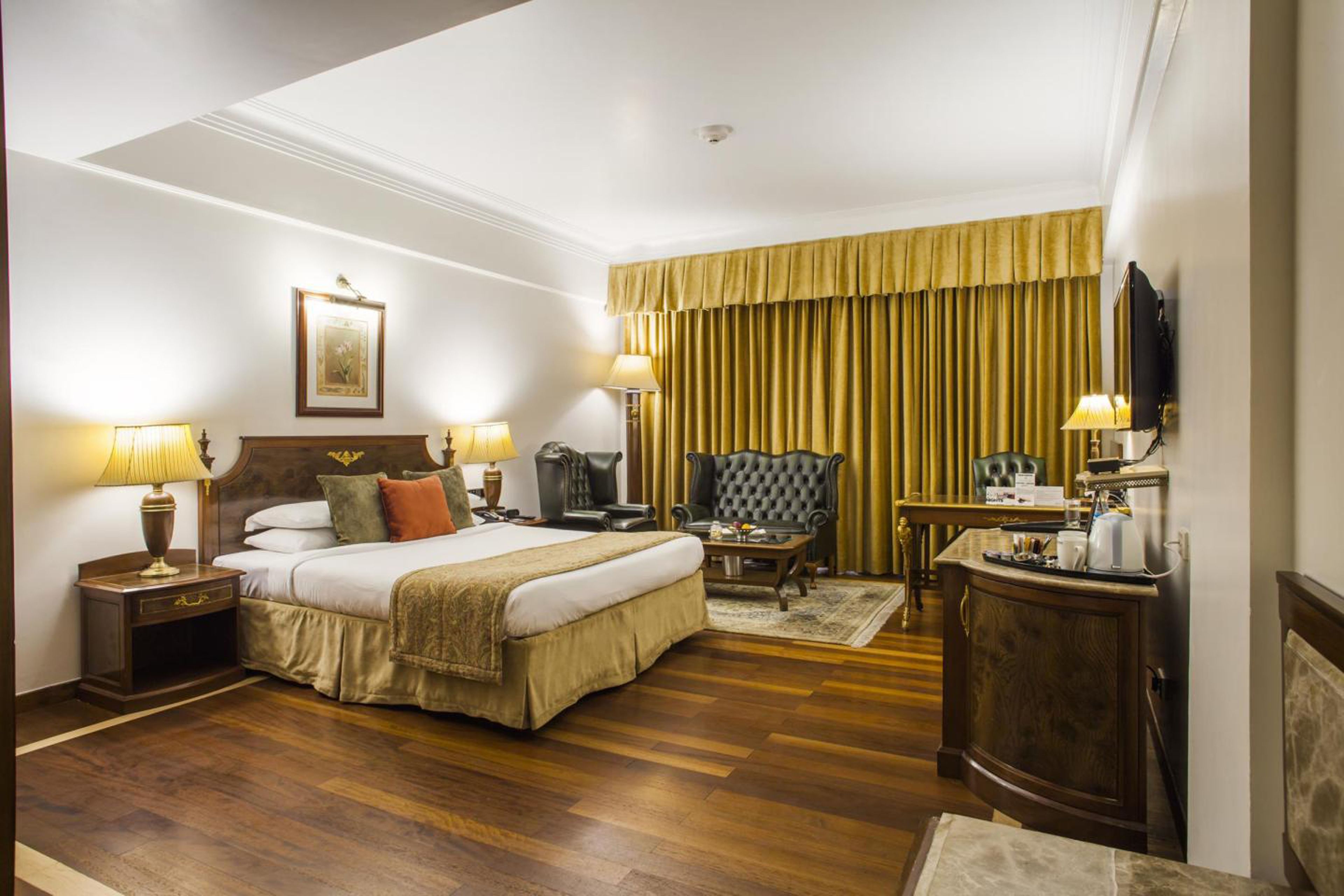 A hotel room with a large bed, two bedside tables with lamps, a chair, a desk with a chair, a television, and a large window with curtains.