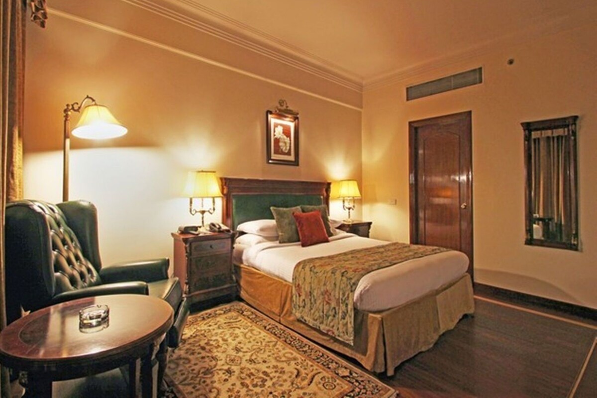 A hotel room with a bed, two bedside lamps, a chair, a small table, a rug, and a door.