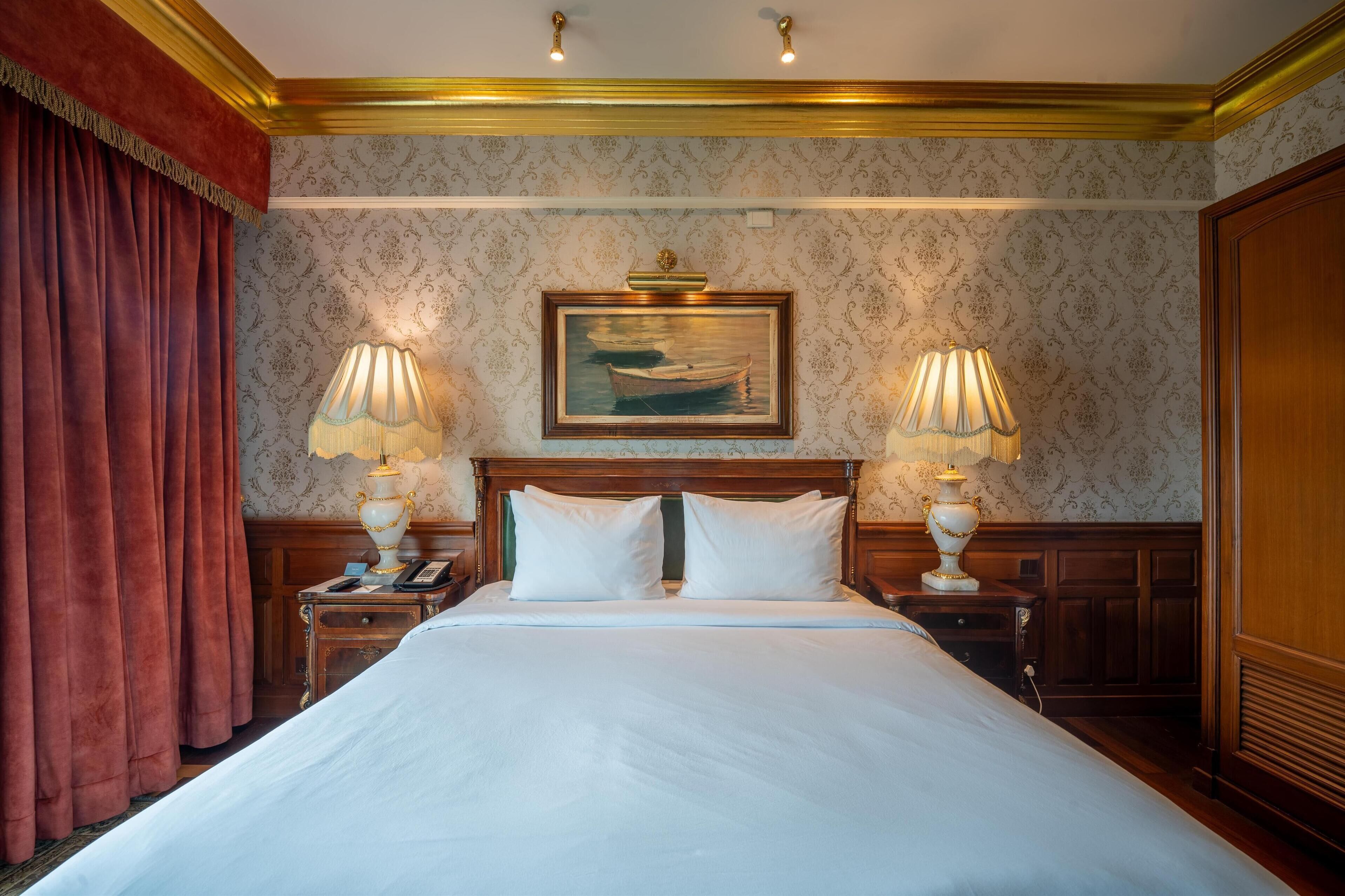 A hotel room with a large bed, two bedside lamps, a painting above the bed, and a wooden headboard.
