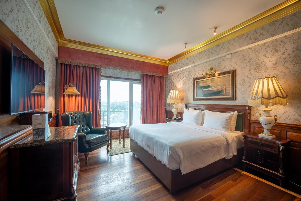 A hotel room with a large bed, two bedside tables, a desk with a chair, a window with curtains, and a painting on the wall.