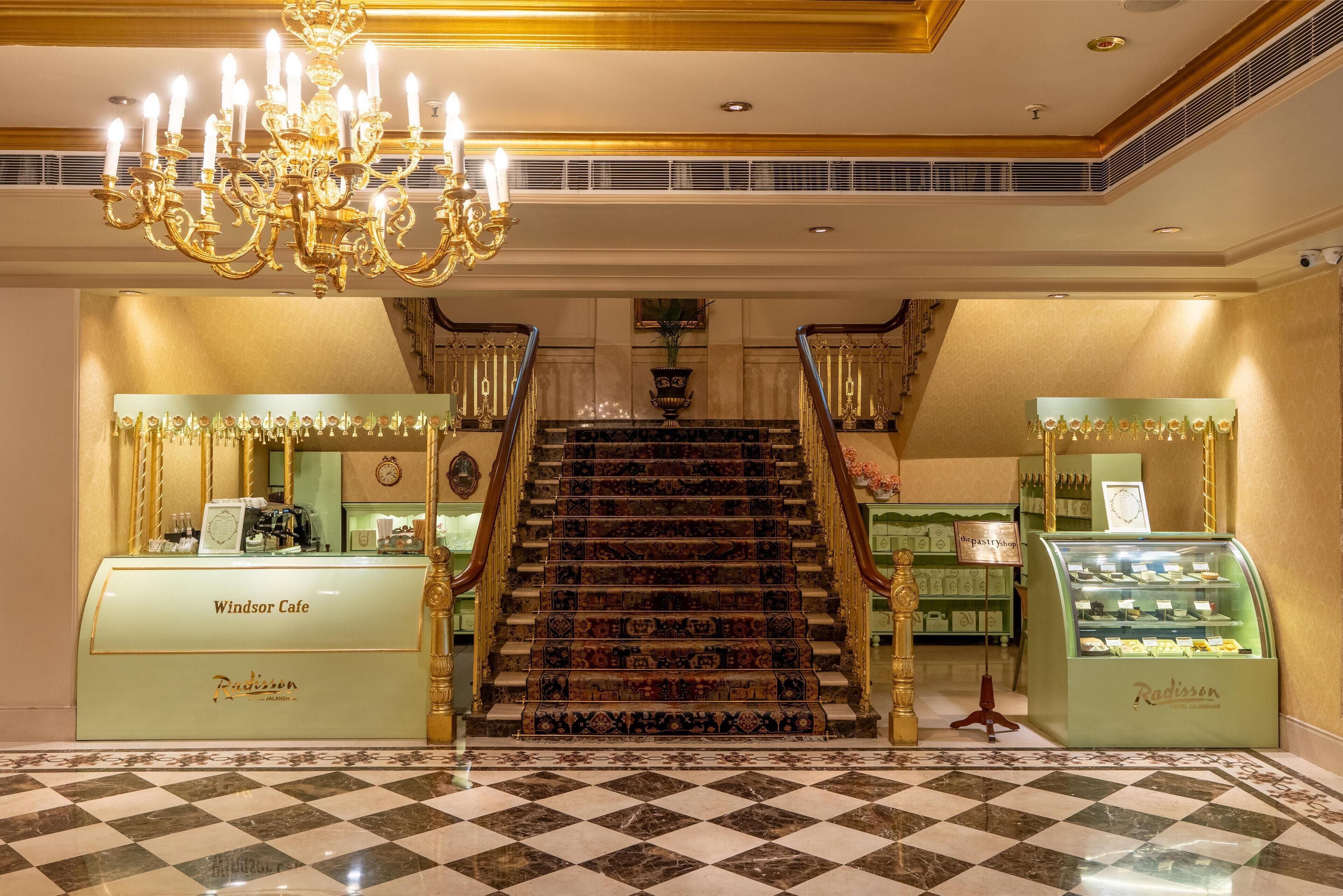 Een statige trap met een kroonluchter, leidend naar een receptiehal met een schachthoefvloer en twee vitrines met de teksten 'Windsor Cafe' en 'Radisson'.