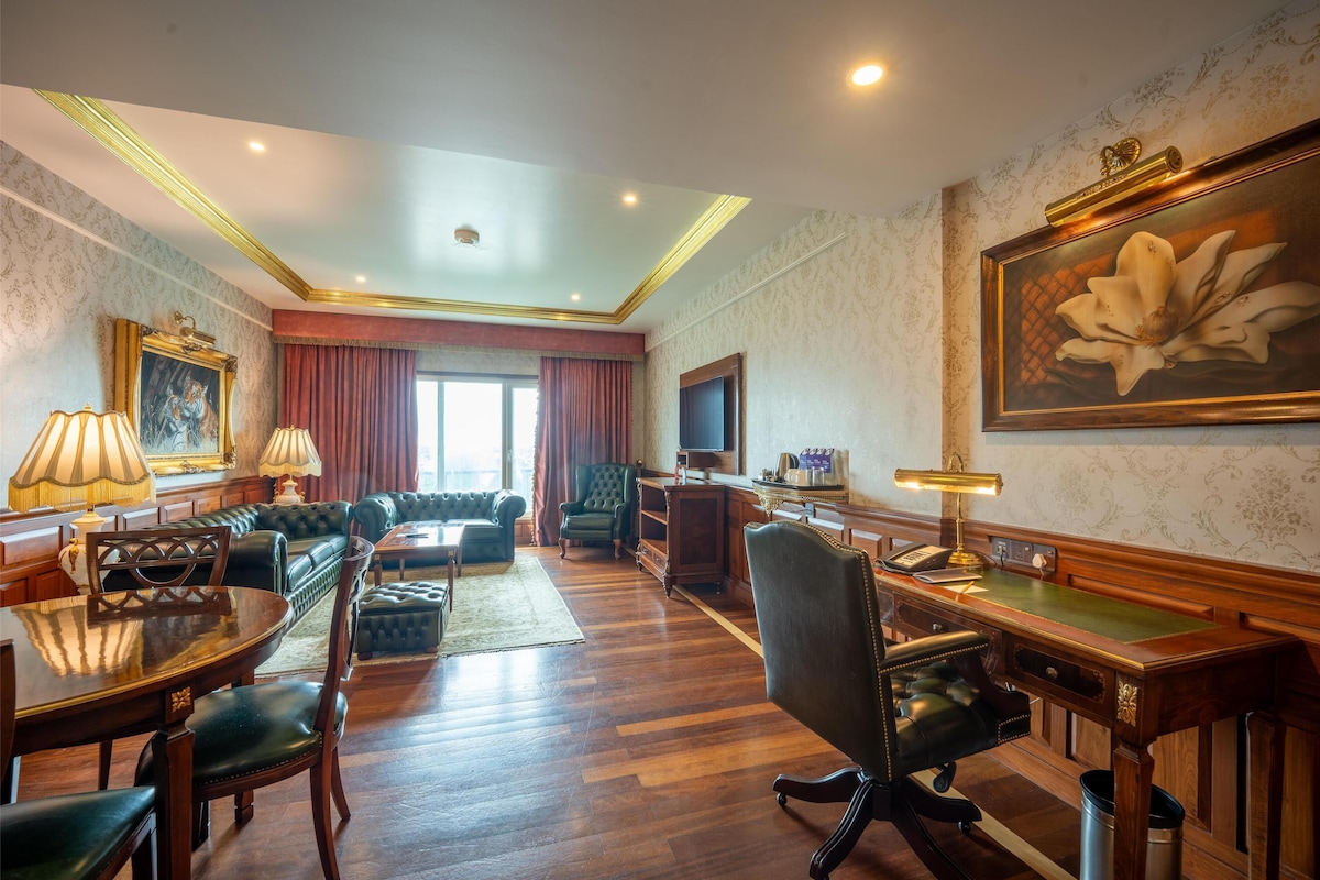 A spacious room with a wooden floor, a green sofa set, a wooden desk with a telephone, and a large framed painting on the wall.