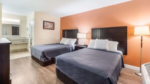 Une chambre d’hôtel avec deux lits, un bureau, une chaise, une lampe et une salle de bain équipée d’un miroir et d’un lavabo.