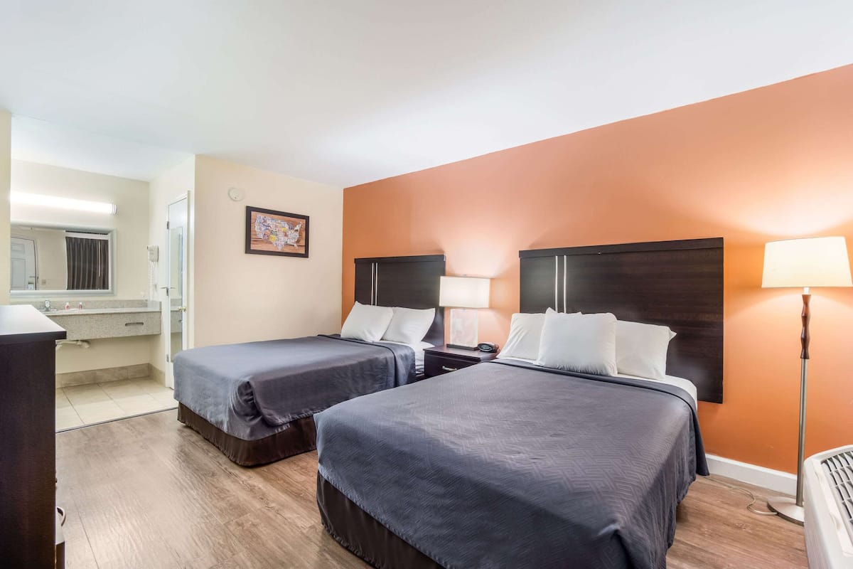 A hotel room with two beds, a desk, a chair, a lamp, and a bathroom with a mirror and sink.