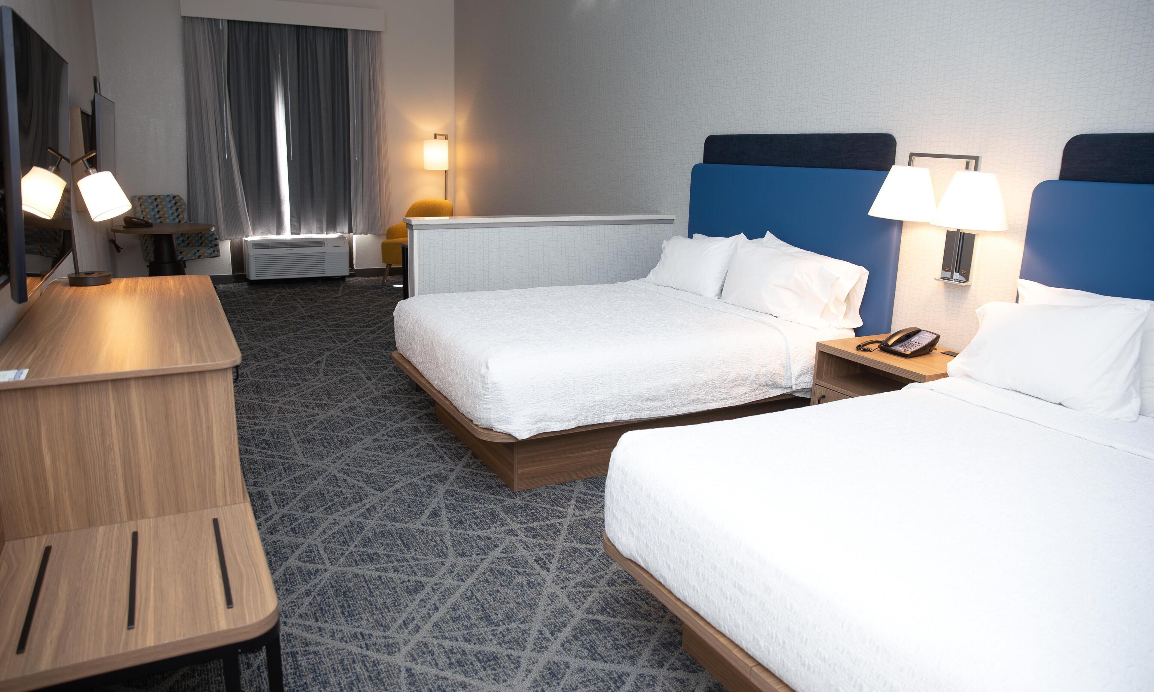 Quarto de hotel com duas camas, uma escrivaninha, uma cadeira, um abajur e um telefone na mesa de cabeceira.