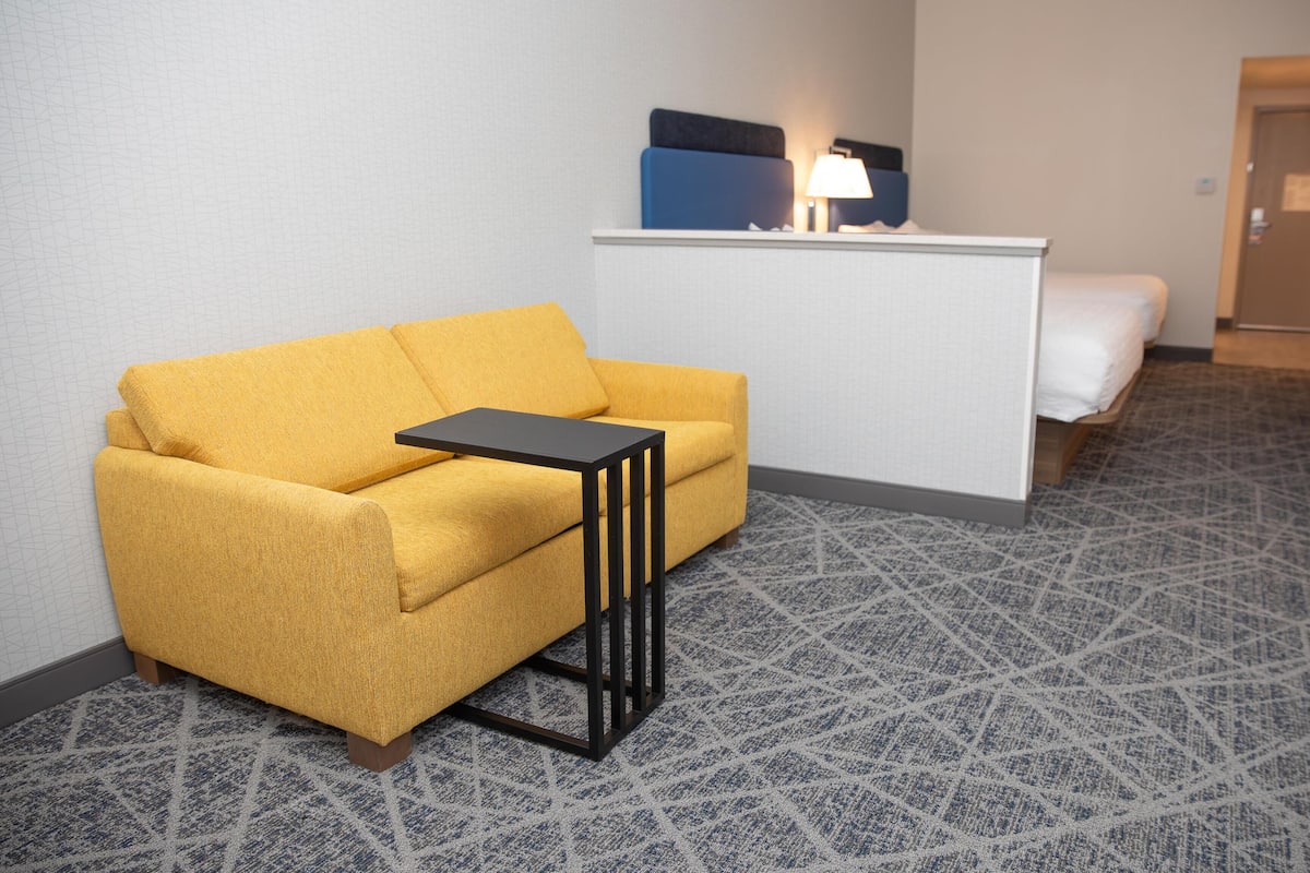 Ein Hotelzimmer mit gelber Couch, einem kleinen schwarzen Tisch, einem weißen Bett und einem Schreibtisch mit Lampe.