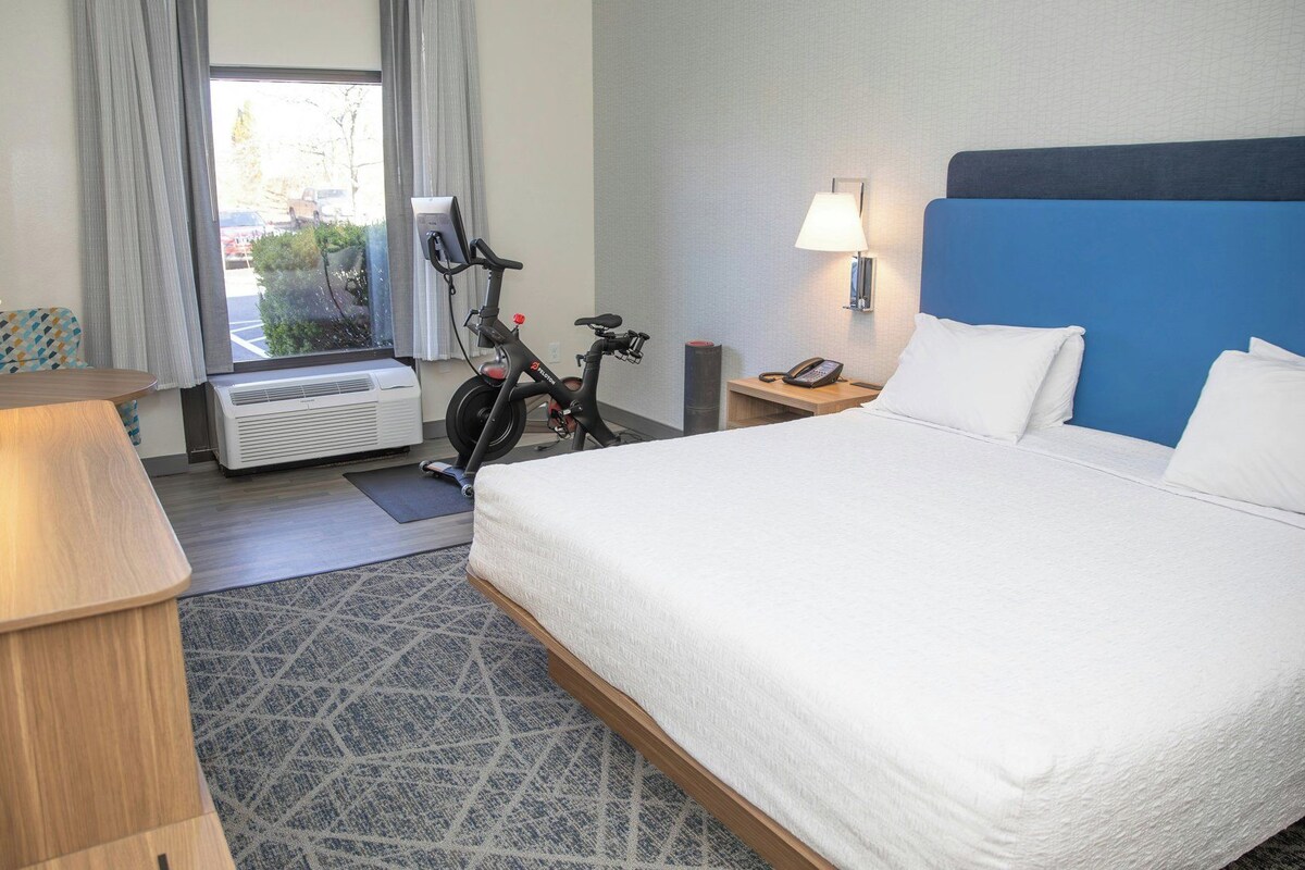 A hotel room with a bed, a desk, a chair, a bicycle, a phone, and a window with curtains.