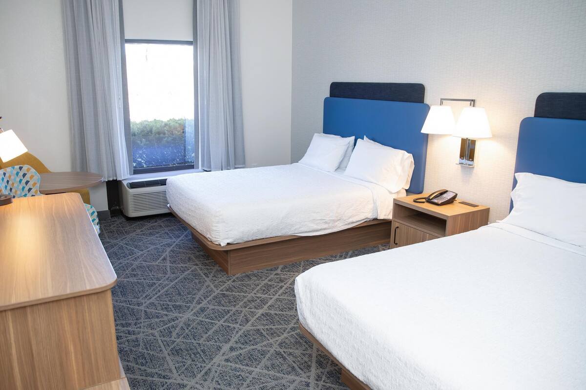 Ein Hotelzimmer mit zwei Betten, einem Schreibtisch, einer Lampe, einem Telefon und einem Fenster mit Vorhängen.
