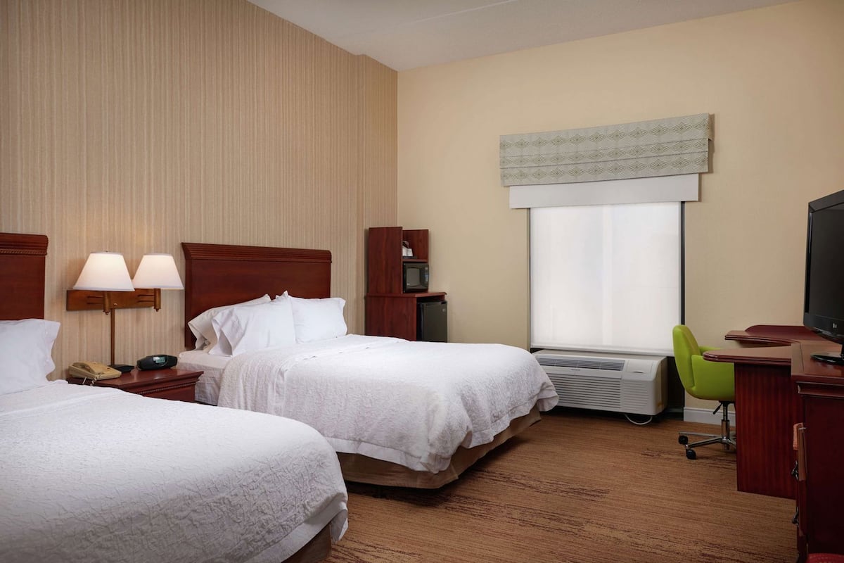 Ein Hotelzimmer mit zwei Betten, einem Schreibtisch mit Stuhl, einem Fernseher, einem Fenster mit Jalousien und einer Klimaanlage.