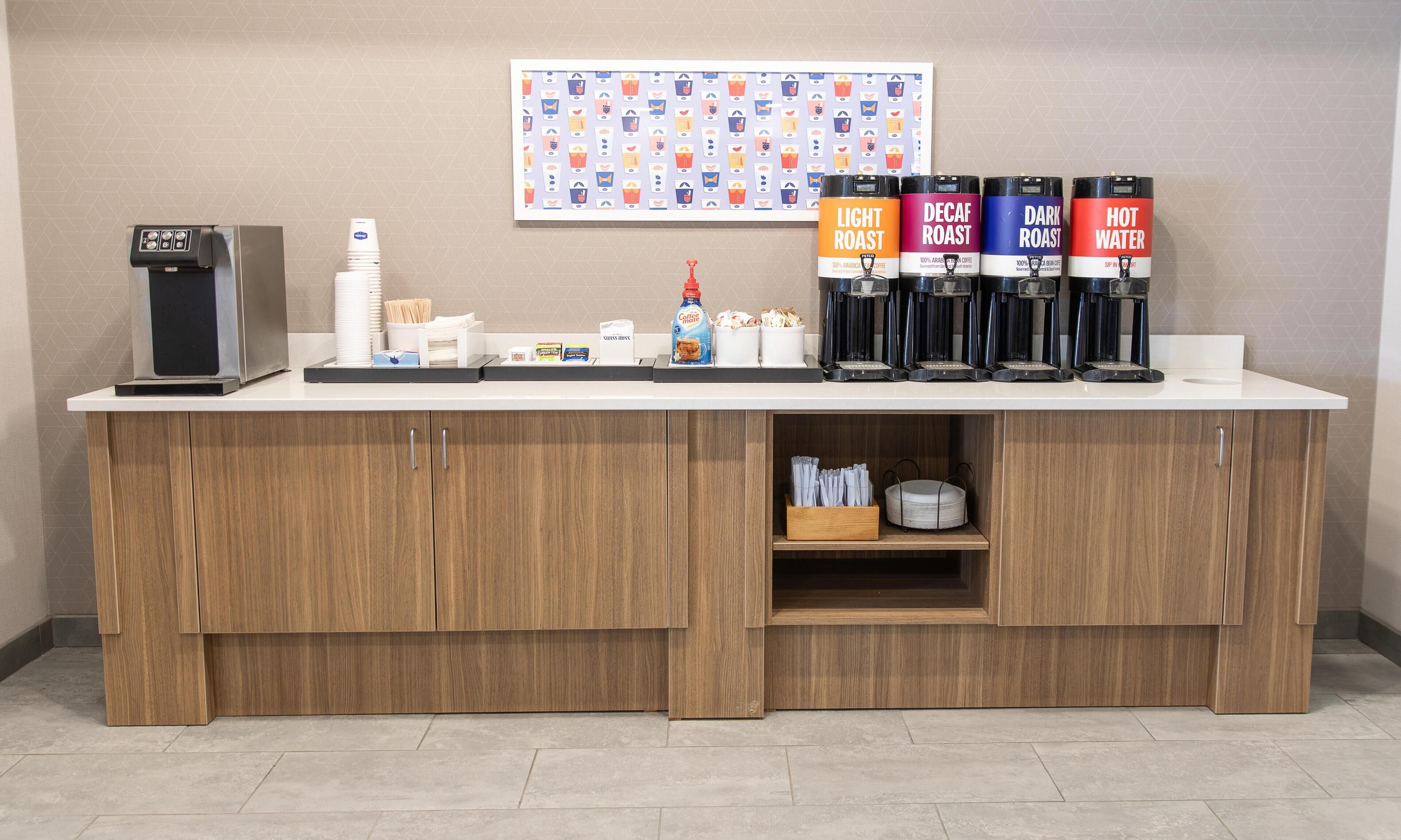 Una estación de café con cuatro dispensadores de café etiquetados como 'Light Roast', 'Decaf Roast', 'Dark Roast' y 'Hot Water'. Hay tazas, una máquina de café y un dispensador de bebidas en la encimera.