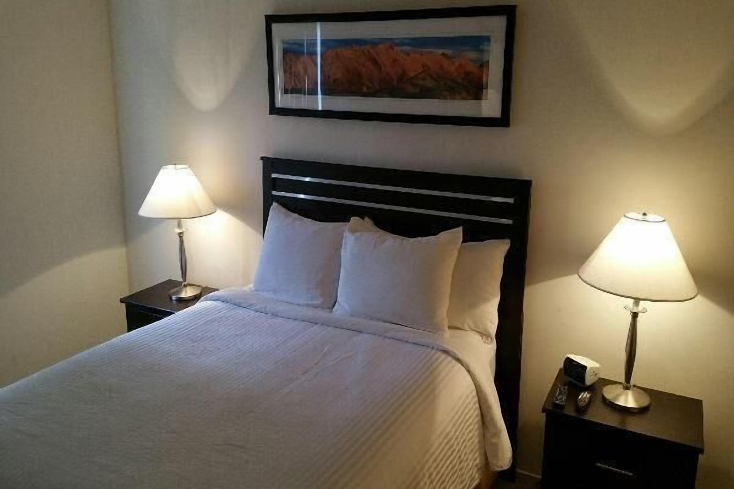 A neatly made bed with white linens, two bedside lamps, a framed picture on the wall, and a small nightstand with a phone and remote control.