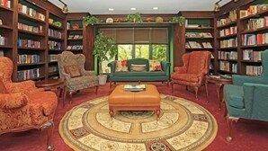 A library with a central sofa, surrounded by bookshelves, a patterned rug, and a window with greenery outside. - Oakwood River Oaks North (Rochester)