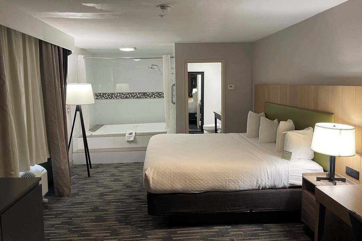 A hotel room with a large bed, bedside table, lamp, and a view of the bathroom.