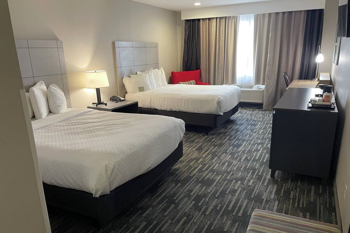 A hotel room with two beds, a desk, a chair, a lamp, and a window with curtains.