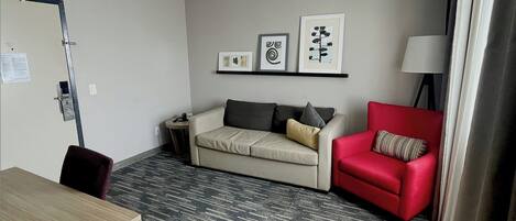 Habitación de hotel con un sofá color beige, un sillón rojo, una mesita auxiliar y una lámpara. Hay tres obras de arte enmarcadas en la pared.