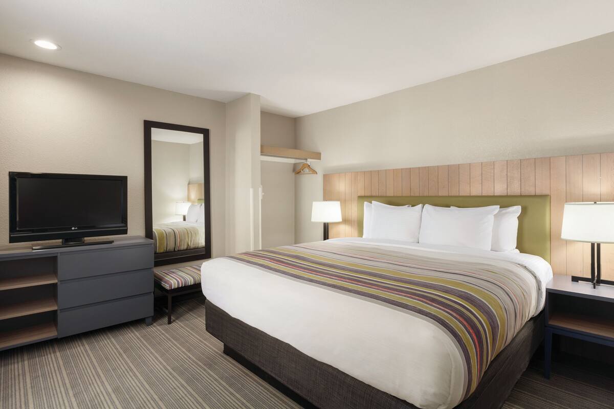 A hotel room with a bed, a TV, a dresser, a bedside table with a lamp, and a mirror on the wall.
