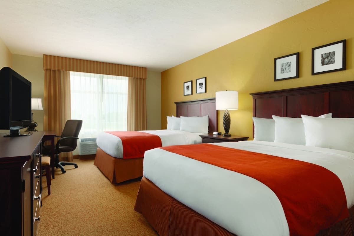 A hotel room with two beds, a desk, a chair, a lamp, and framed pictures on the wall.
