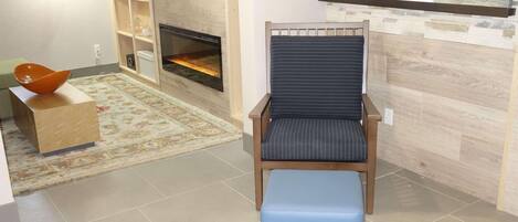 A living room with a fireplace, a blue ottoman, and a wooden chair with a striped cushion.