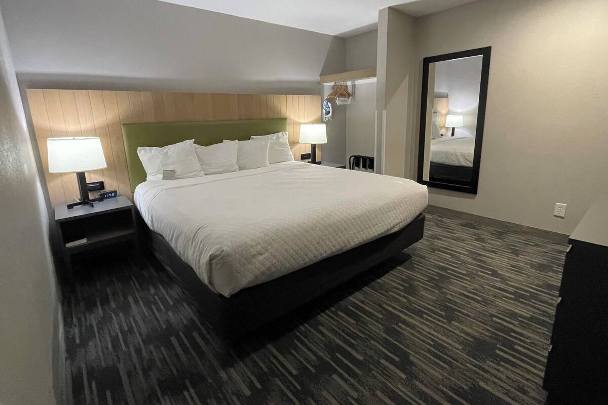 A hotel room with a large bed, two bedside tables with lamps, a wooden headboard, a carpeted floor, a wall-mounted mirror, and a clock.