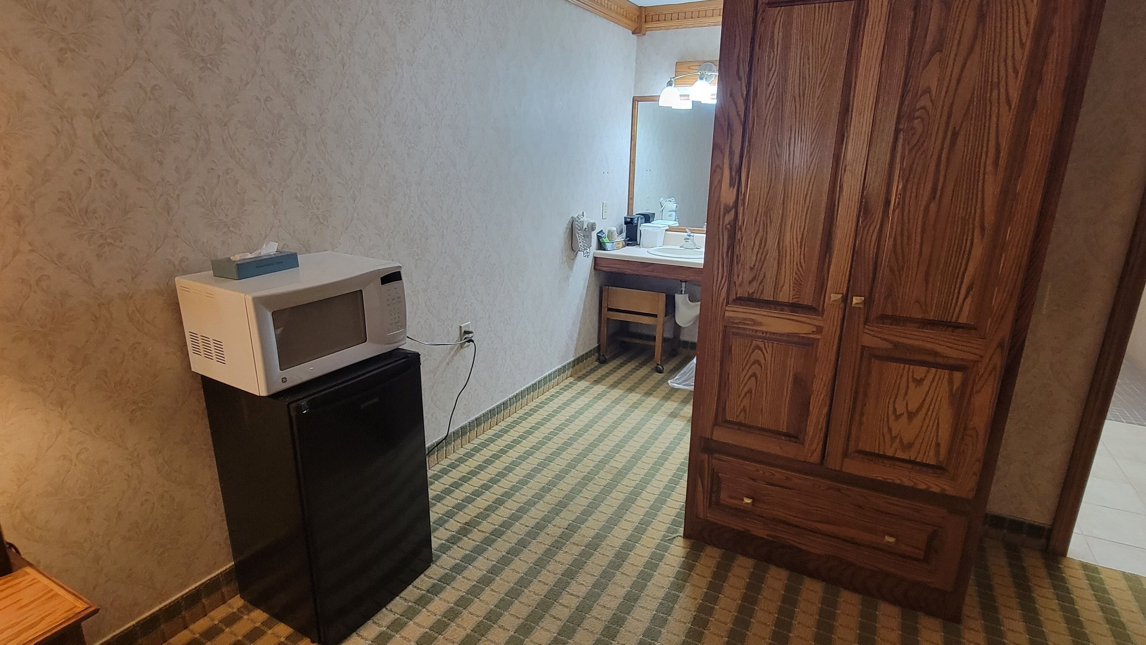 Habitación compacta con microondas sobre un refrigerador negro, un armario de madera y un escritorio pequeño con un espejo.