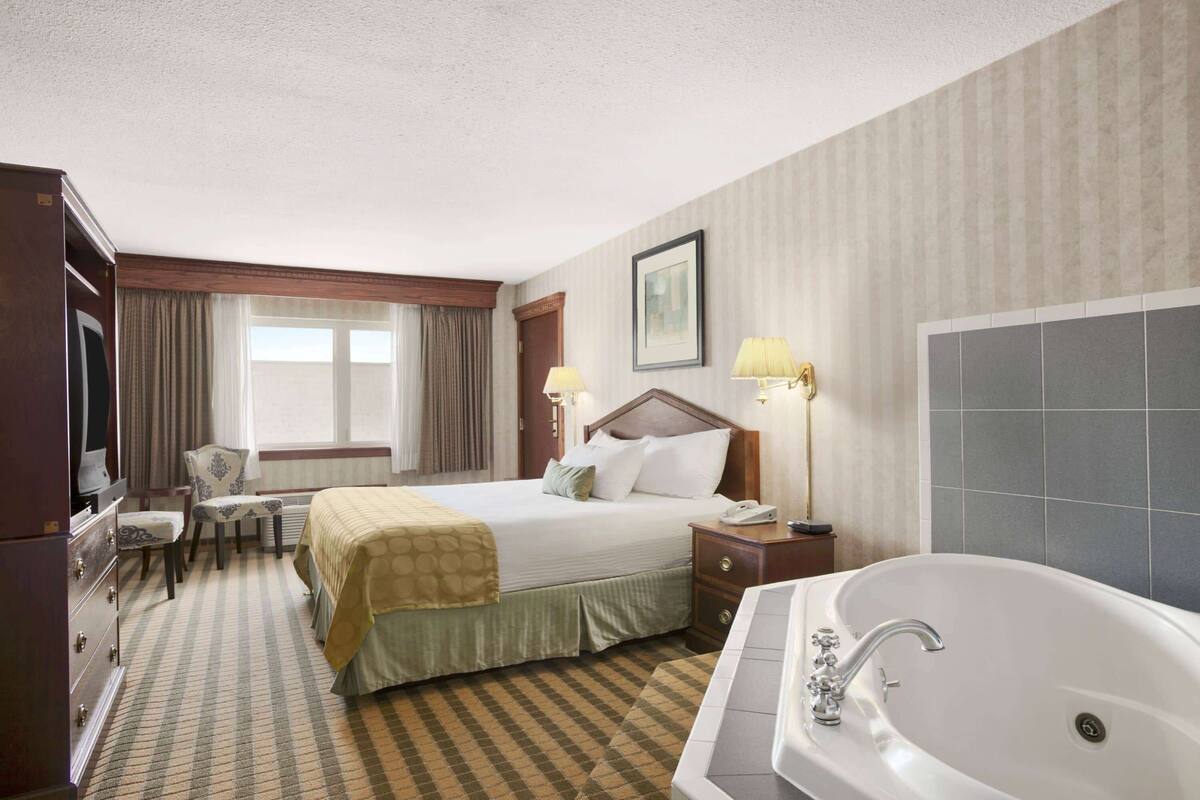 A hotel room with a large bed, a desk, a chair, a TV, a desk lamp, a bedside table, a telephone, and a bathtub.
