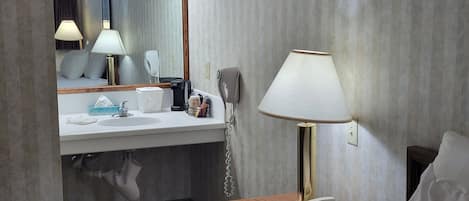 A hotel bathroom with a sink, mirror, towel rack, and a bedside table with a lamp and a phone.