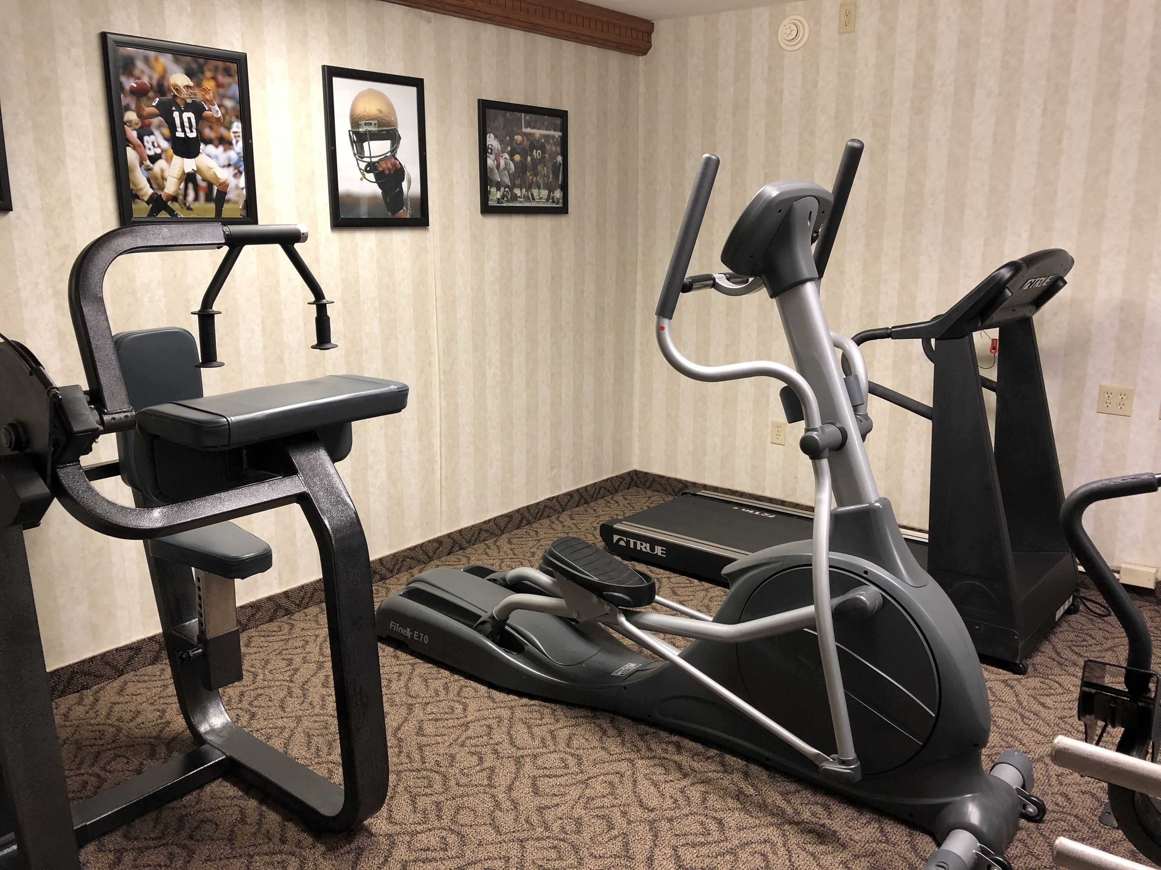 Een fitnessruimte thuis met een elliptische trainer, een hometrainer en een bankdrukapparaat. Op de muur hangen ingelijste sportmemorabilia.